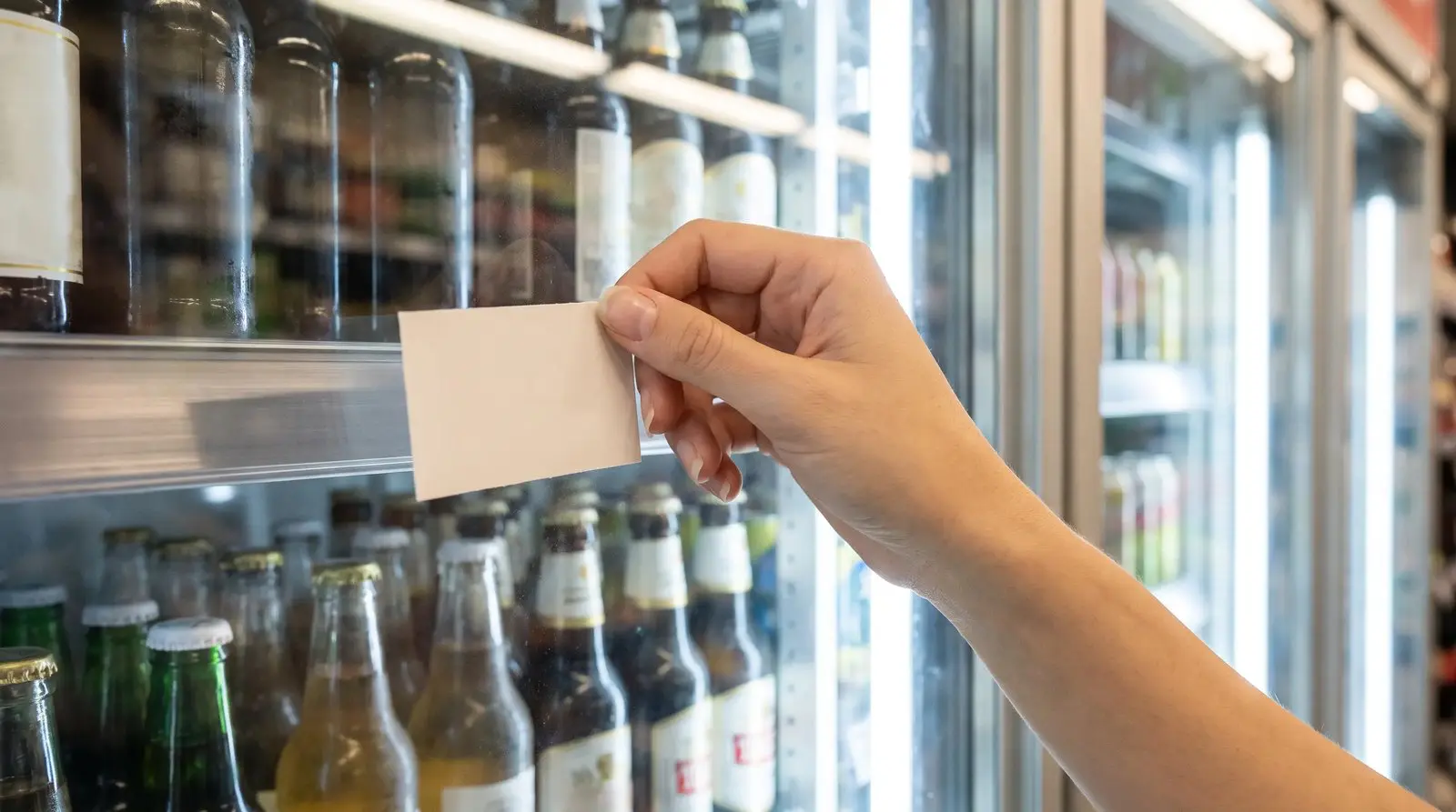 Hand placing a price tag on the shelf edge of a stocked beverage cooler in a California store